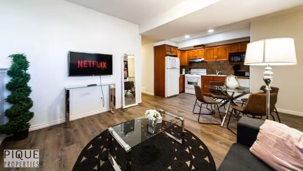 Una sala de estar moderna con una mesa de centro de vidrio, un televisor montado en la pared y una cocina visible a través de una puerta abierta.