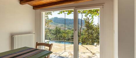 Ein Zimmer mit Holzbalkendecke, einem Tisch mit farbenfroher Webdecke, einem Stuhl und einer Glastür, die zu einem Außenbereich mit Pool und Hügellandschaft führt.