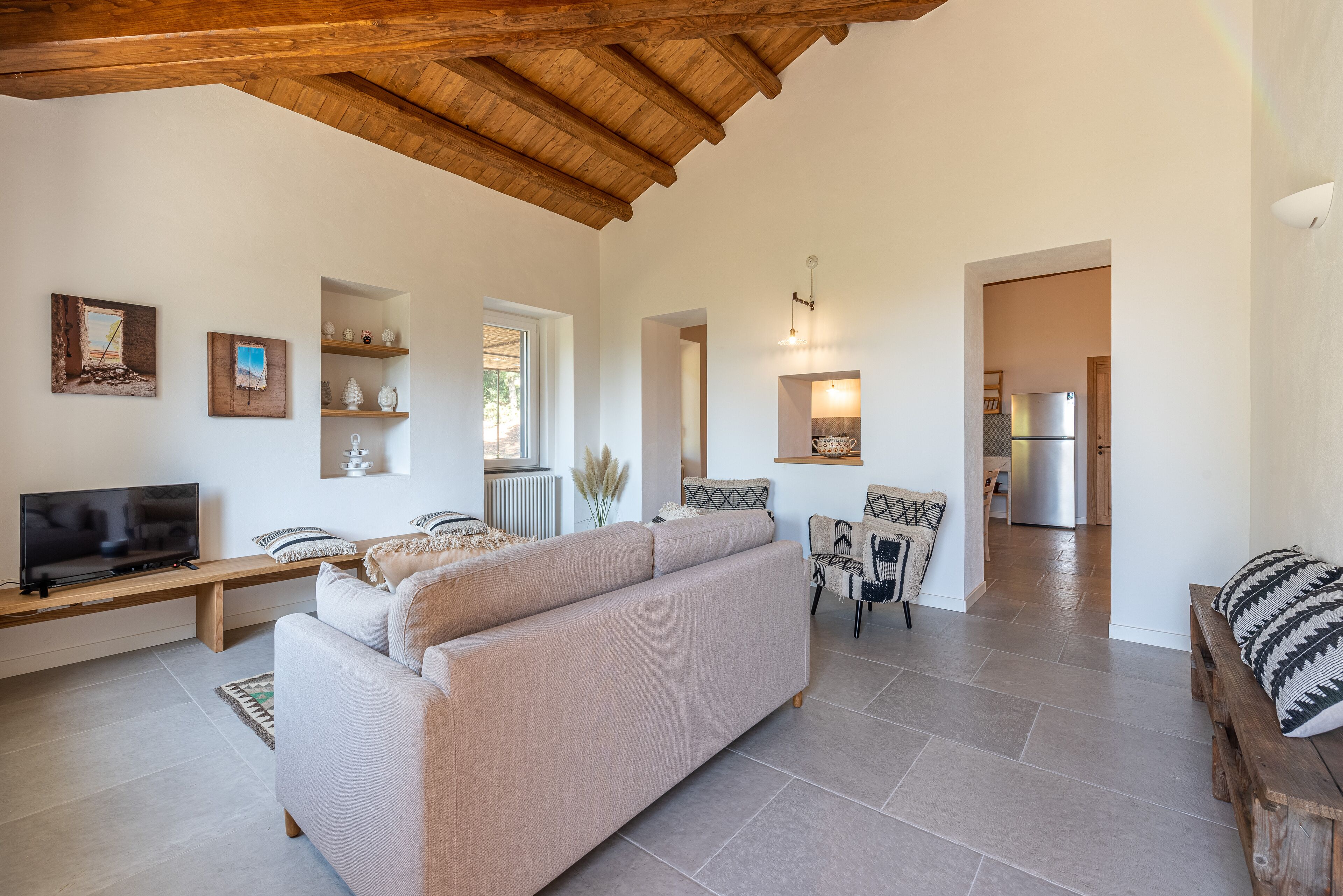 A spacious living room with a large sofa, wooden beams on the ceiling, a television, and a kitchen in the background.