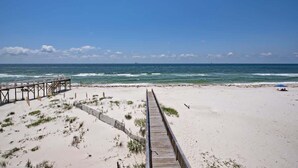Ein Strand mit einem hölzernen Steg, der zum Meer führt, sandigen Dünen und einem strahlend blauen Himmel.