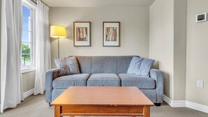 Ein Wohnzimmer mit grauem Sofa, zwei gerahmten Kunstwerken, einer Stehlampe und einem Holztisch.
