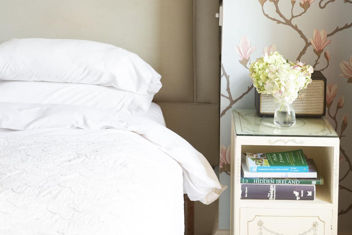 A bedroom with a bed, a nightstand with books and a vase, and floral wallpaper.