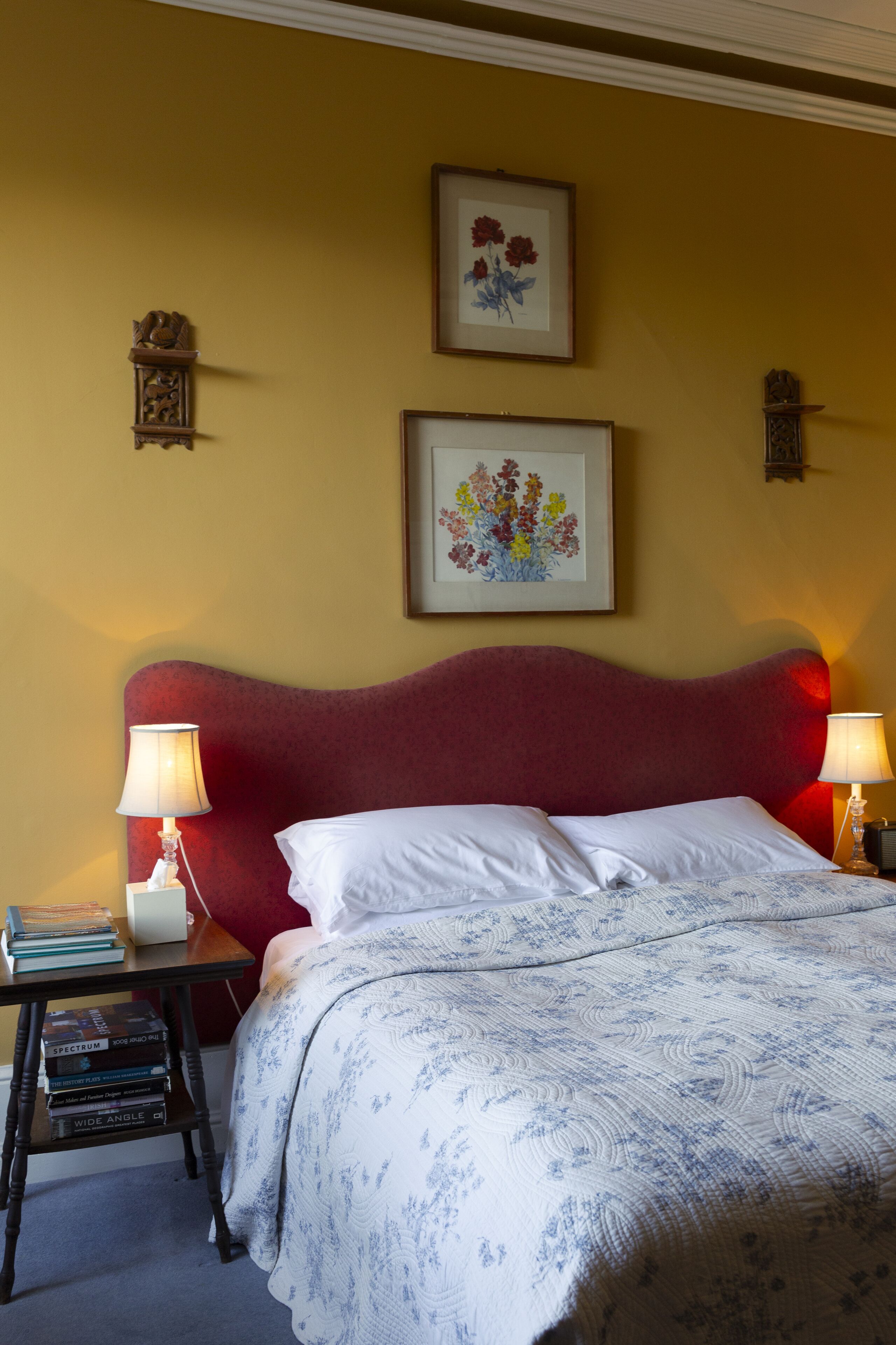 Una cama bien hecha con un cabecero rojo, dos lámparas de noche y una obra de arte con flores enmarcada encima.