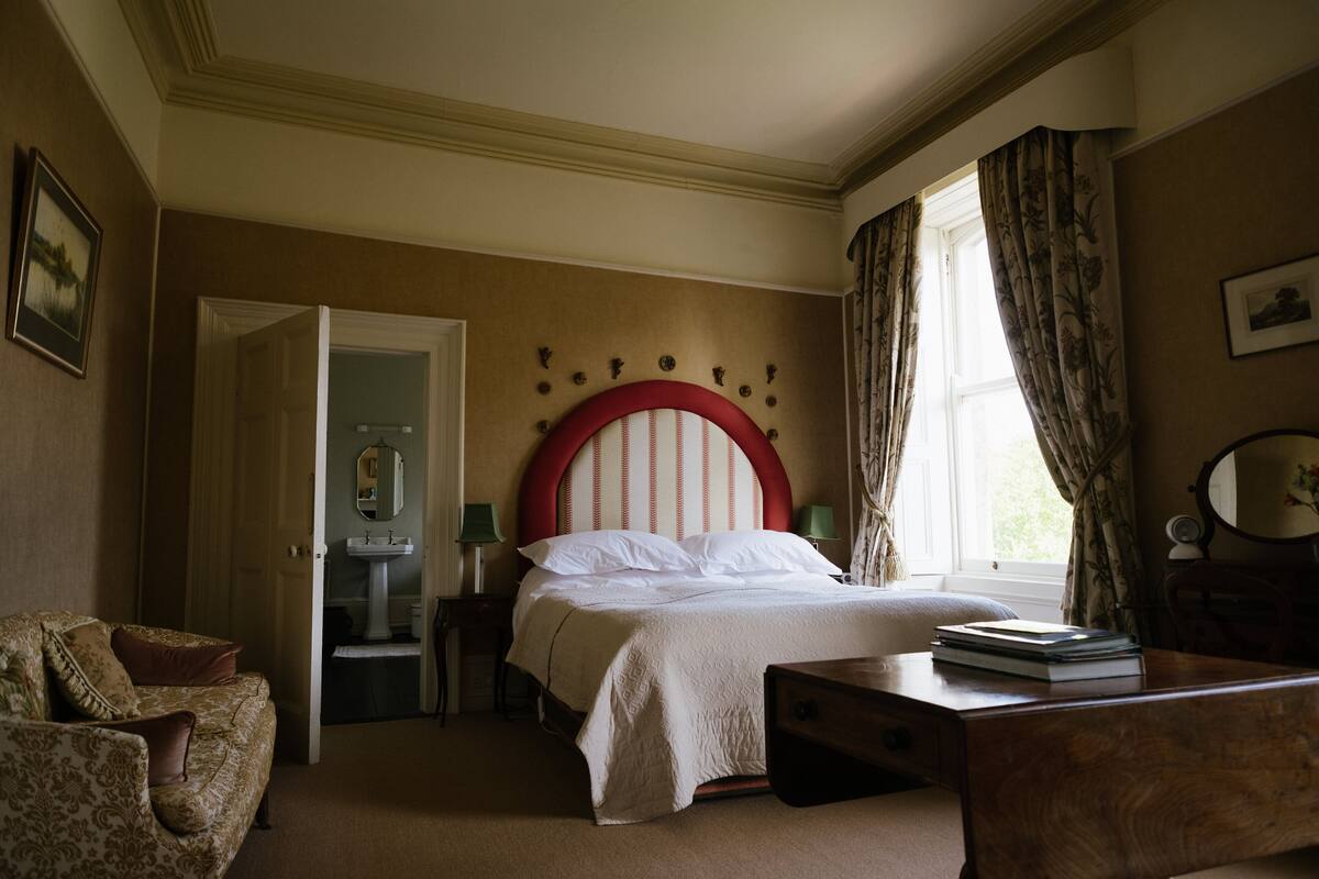 A bedroom with a bed, a wooden nightstand, a dresser, a chair, and a bathroom visible through an open door.