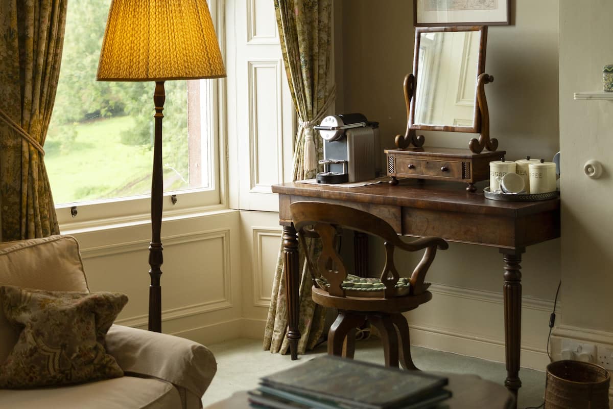 A room with a window, a desk with a lamp, a chair, a sofa, and a small table with books.