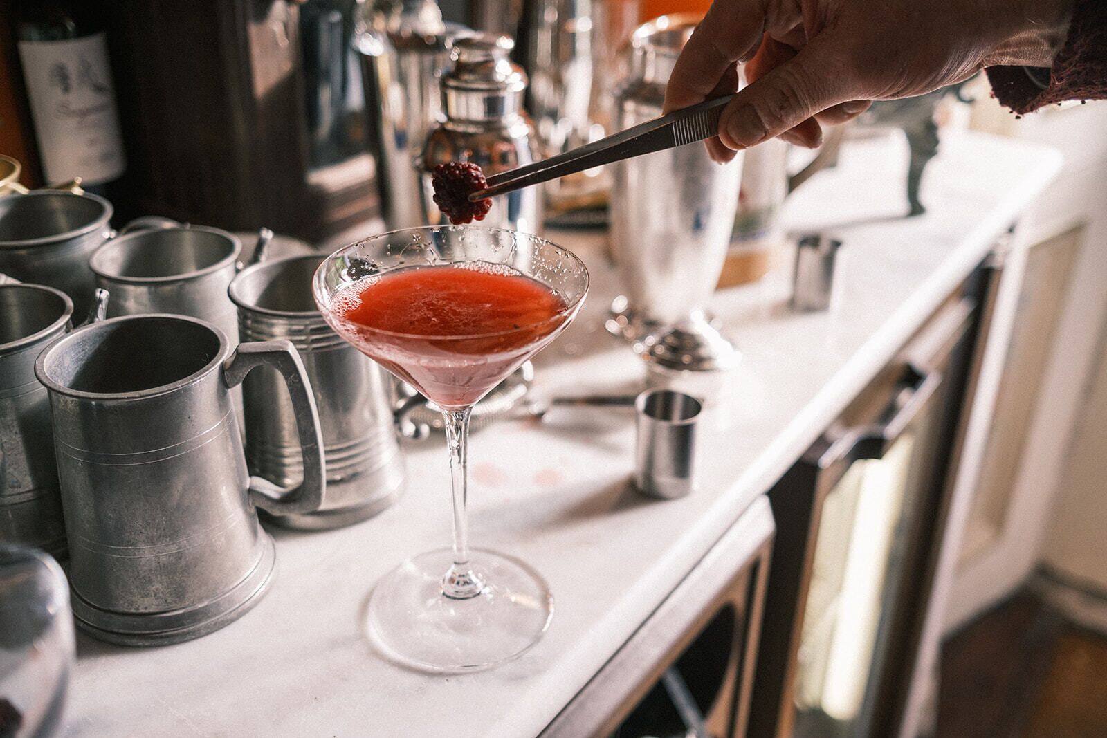 Una copa de martini con una bebida roja, una jarra de metal y una cuchara con una baya encima.