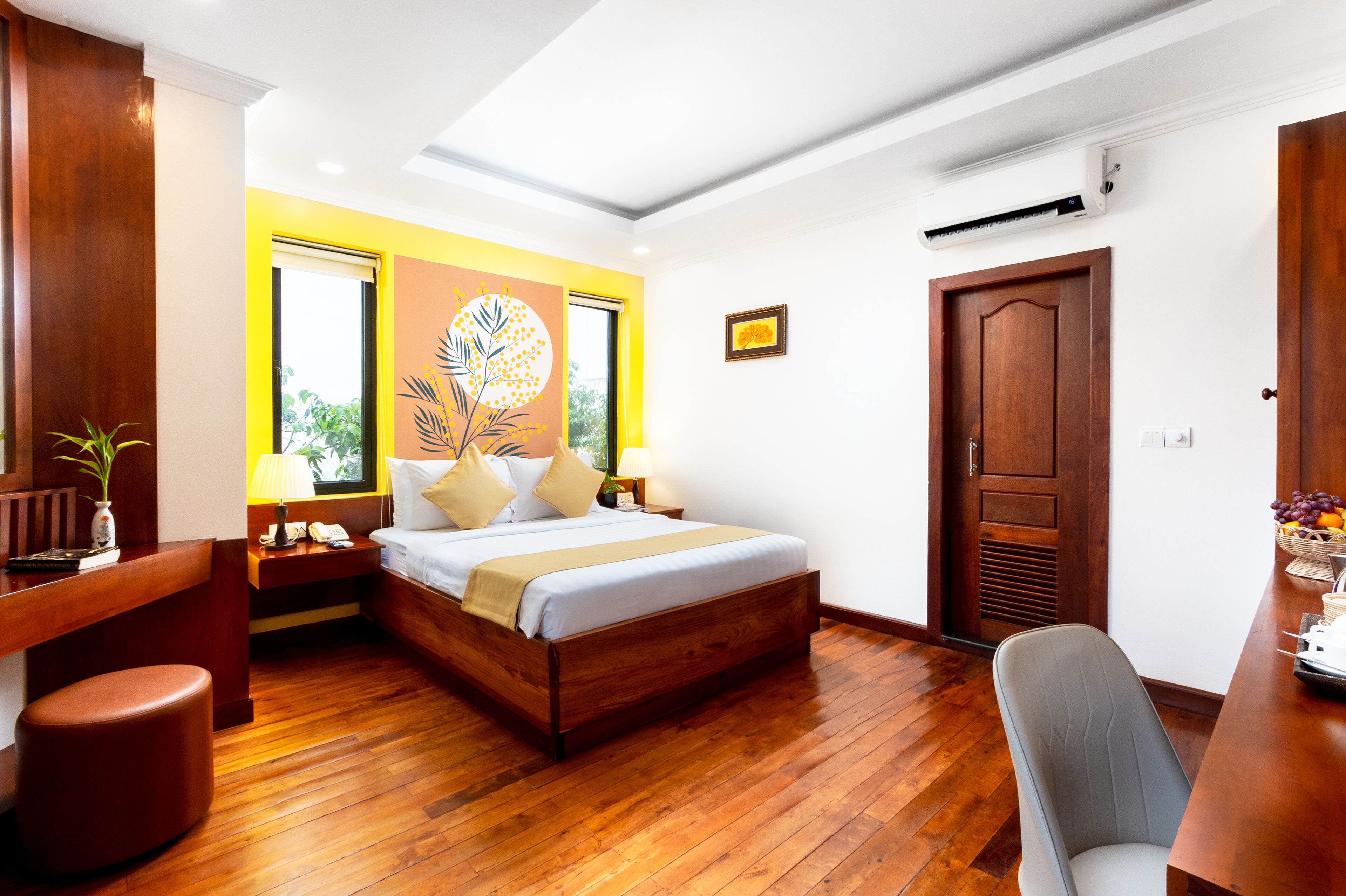 A hotel room with a wooden bed, a desk, a chair, a nightstand, a lamp, a wall art piece, and a door.