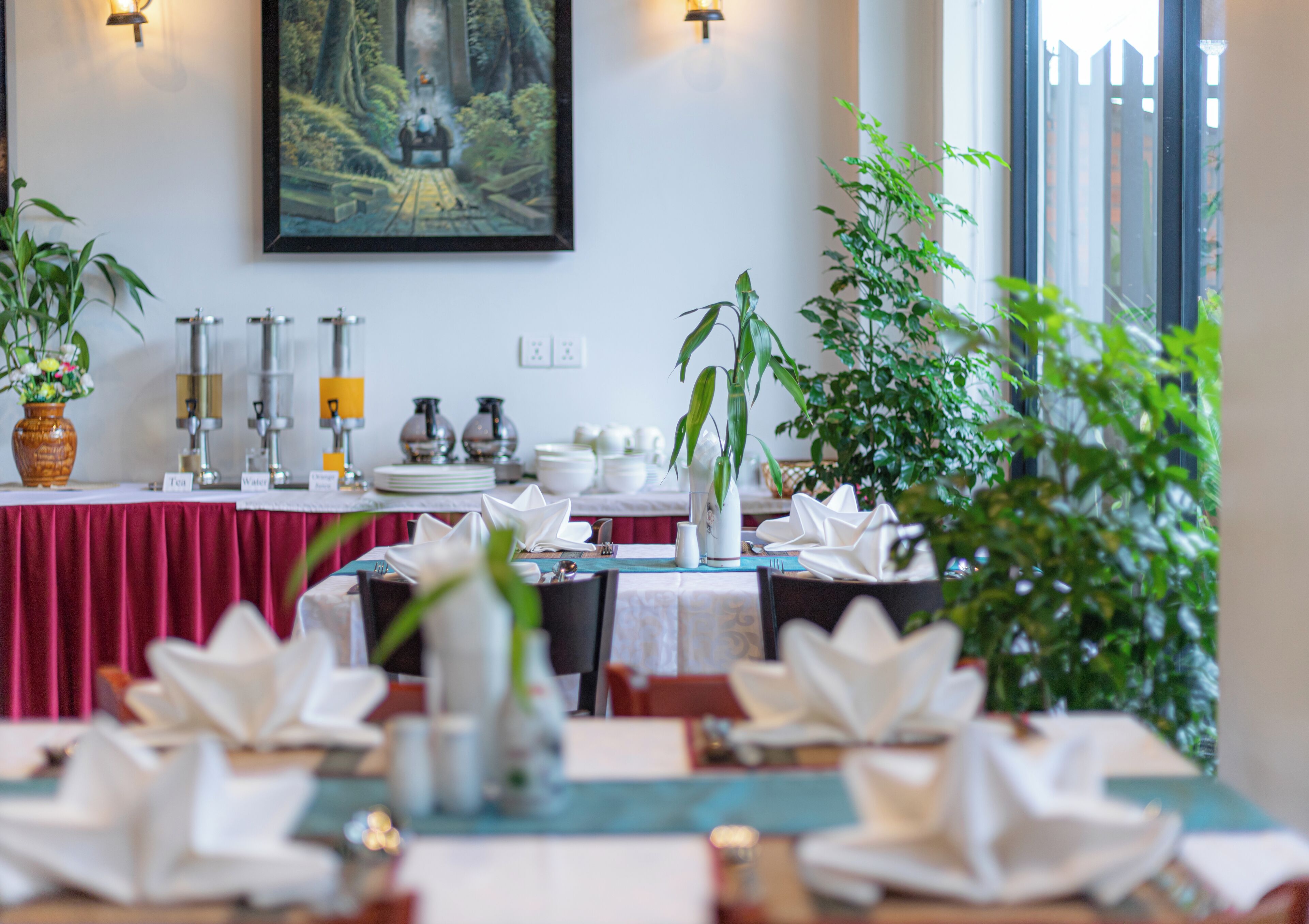 A dining area with tables set for service, a painting on the wall, and potted plants.