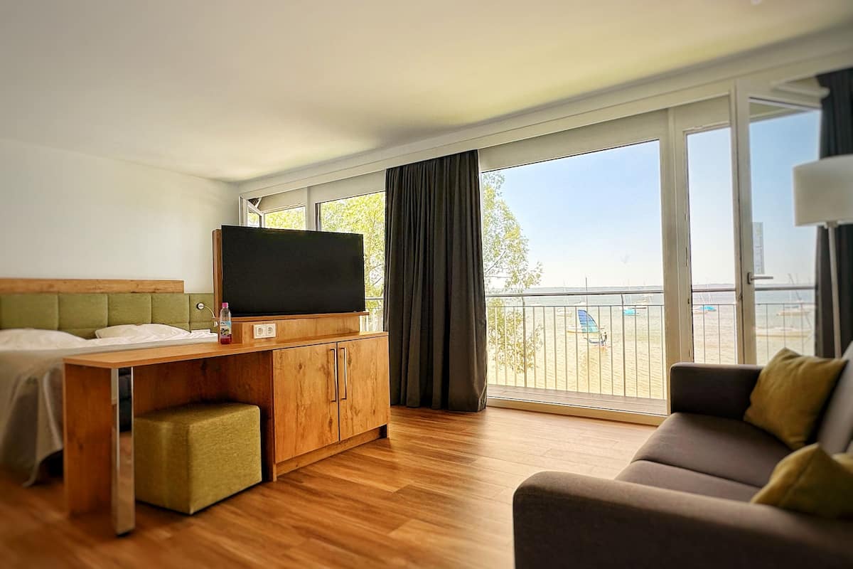 A hotel room with a wooden desk, a television, a sofa, and a balcony with a view of the beach.