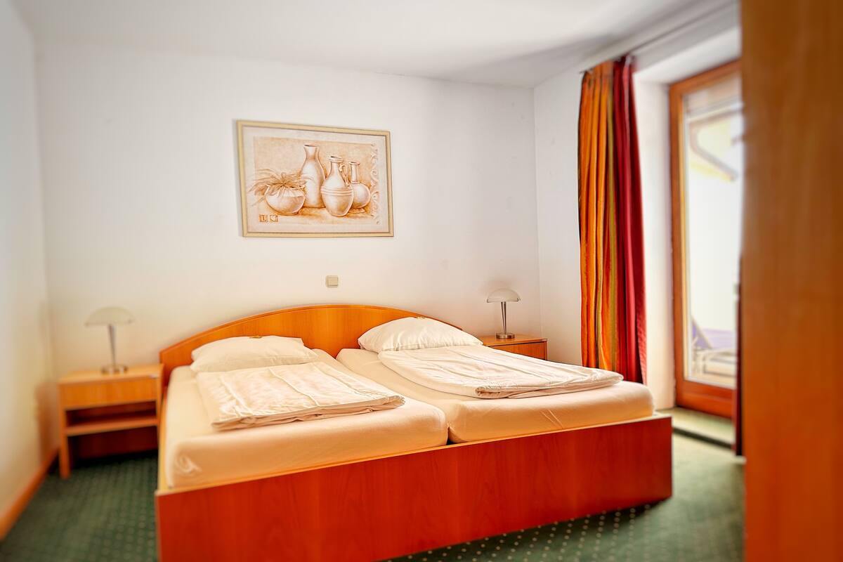 A hotel room with a wooden bed, two bedside tables with lamps, a framed picture on the wall, and a window with curtains.