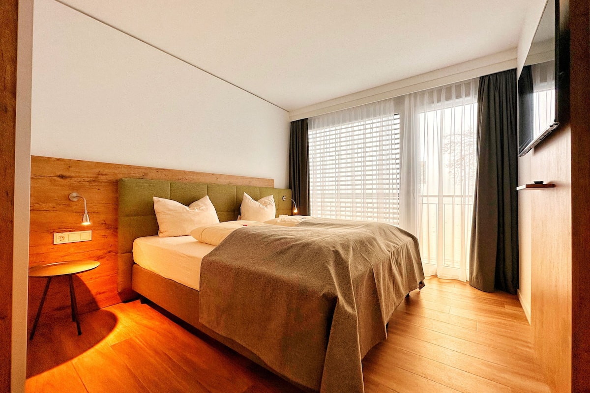 A modern bedroom with a wooden headboard, a bed with a beige bedspread, a bedside lamp, and a large window with curtains.