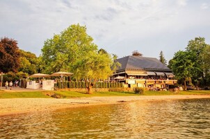 Um edifício à beira do lago com área de estar ao ar livre, rodeado por árvores e uma área de areia.