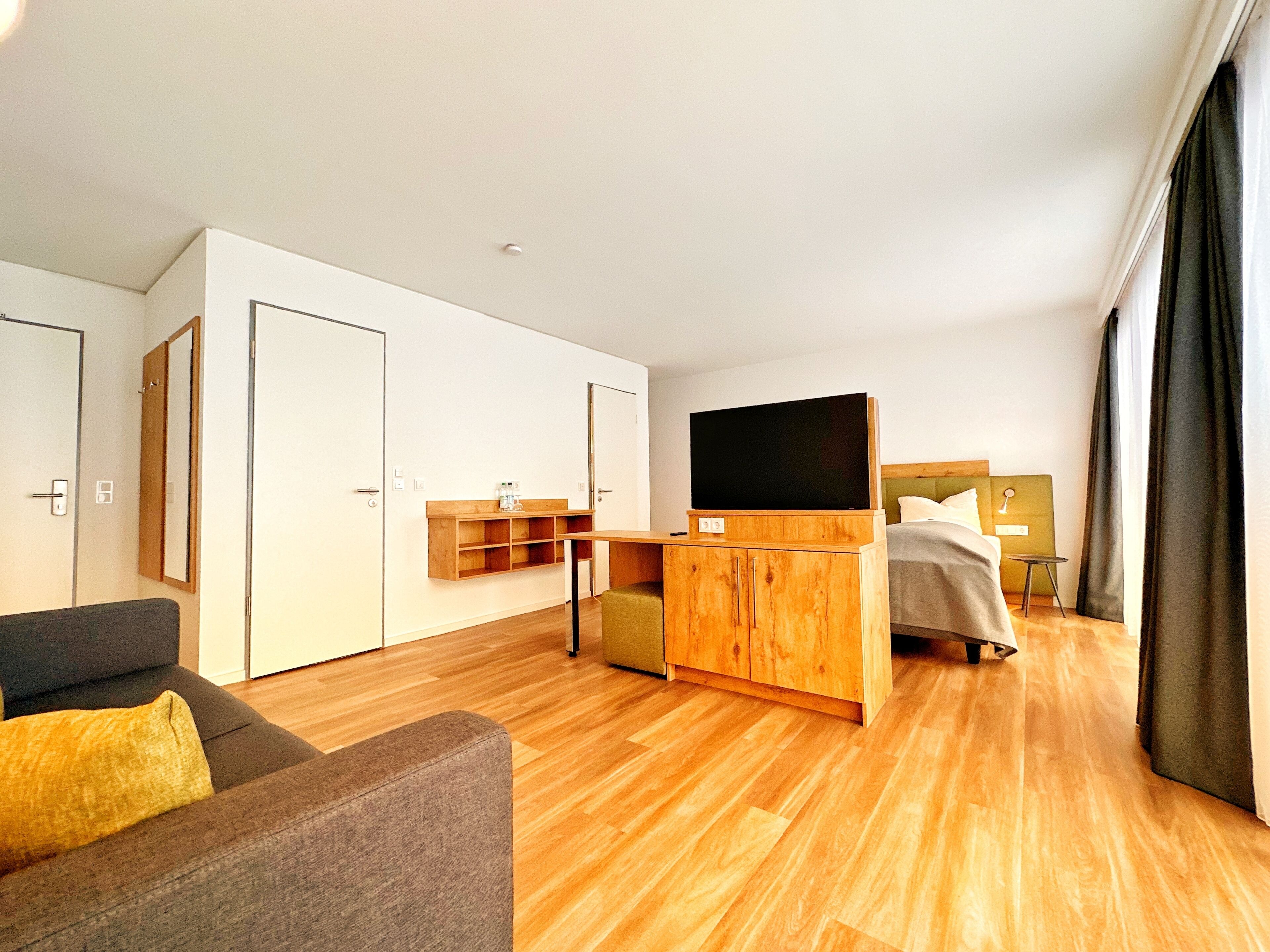 A modern hotel room with a wooden floor, a sofa, a bed, a TV, and a small table.