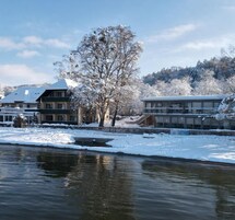 Cena de inverno com um rio, árvores cobertas de neve e edifícios ao longo da margem.