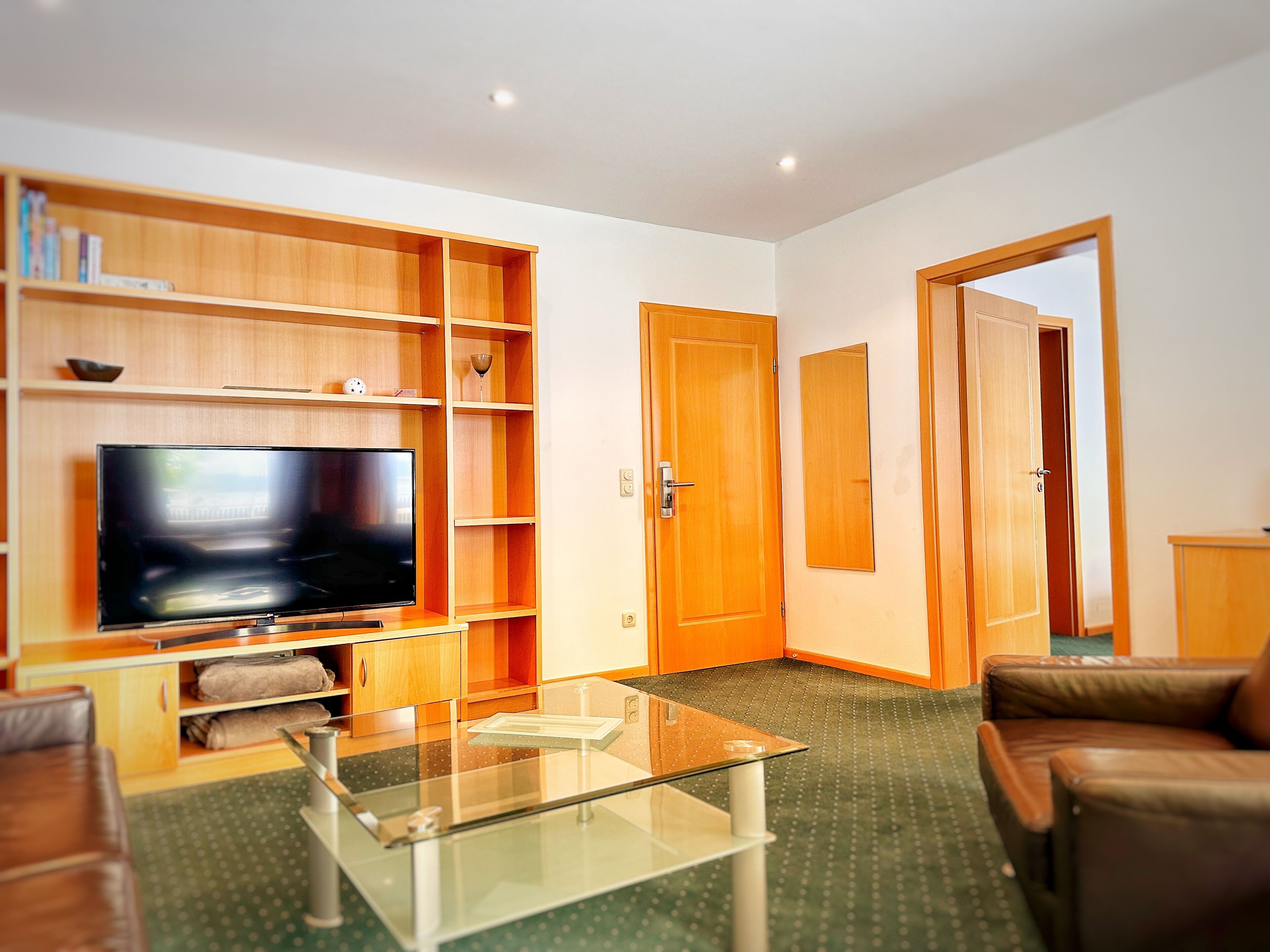 A living room with a flat-screen TV, wooden shelving, a glass coffee table, and a leather sofa.