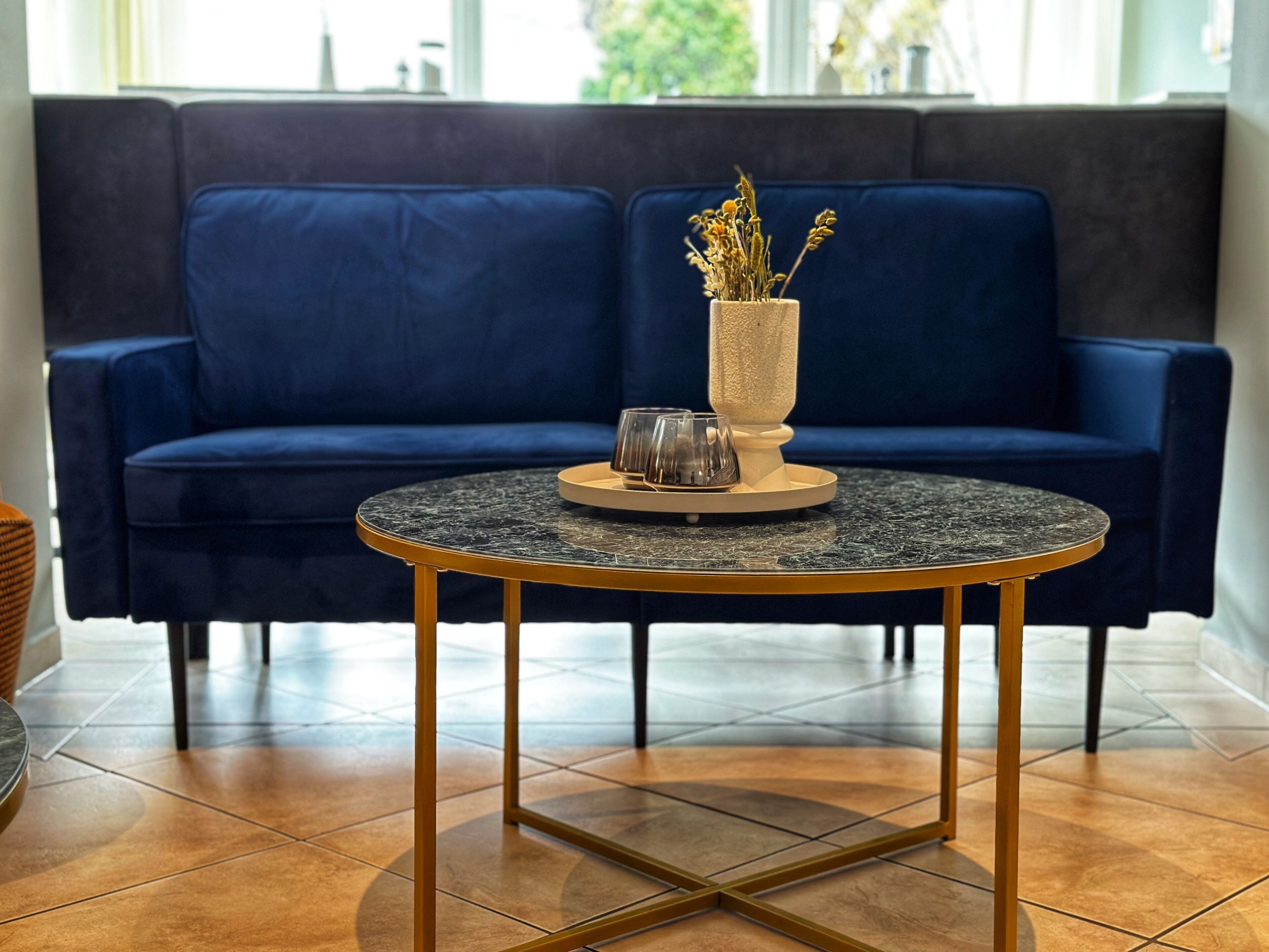 En moderne stue med en mørkeblå sofa, et rundt sofabord med marmorplade og en vase med tørrede blomster.