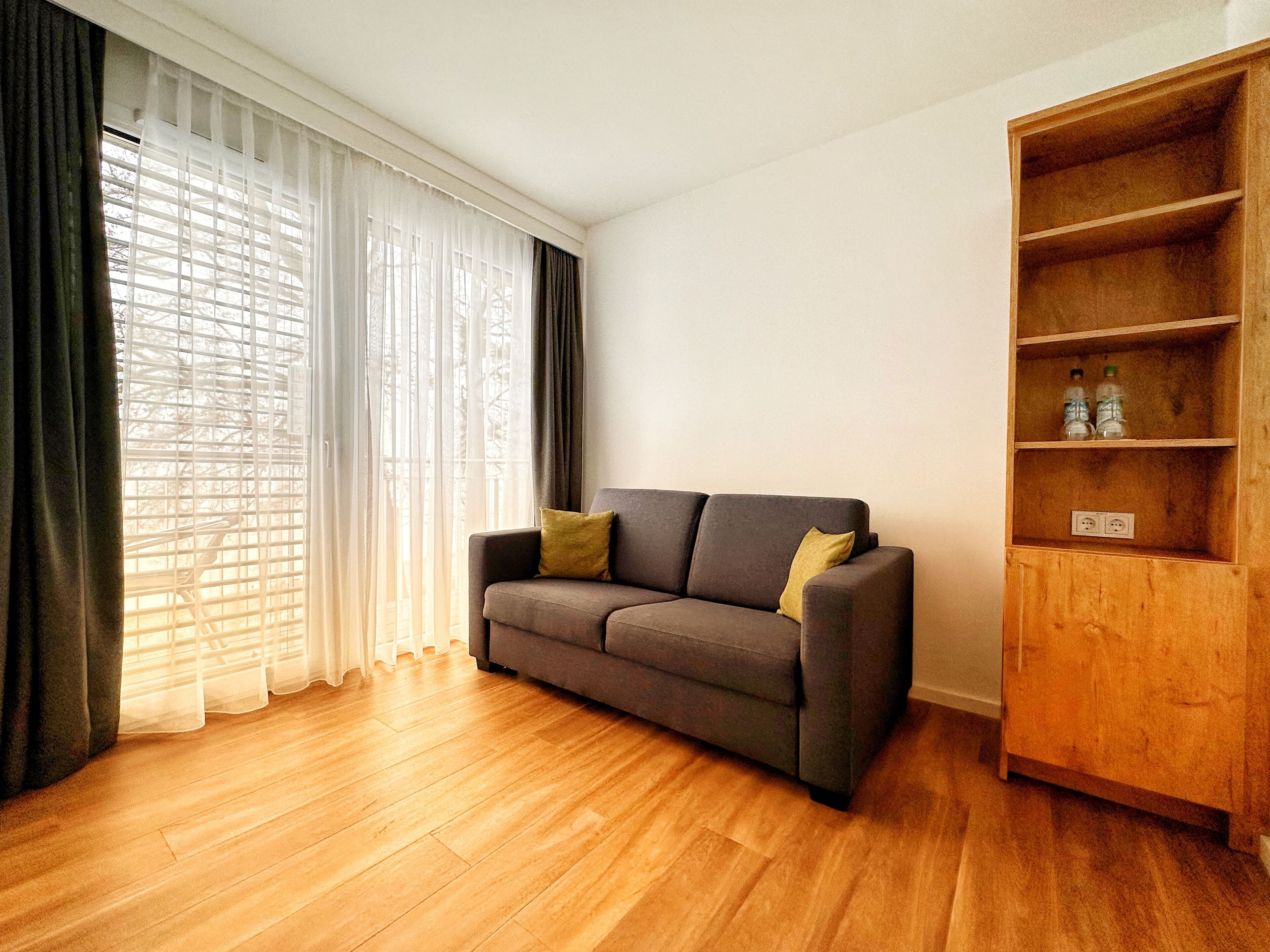 Una sala de estar con un sofá gris, una estantería de madera y un ventanal con cortinas traslúcidas.
