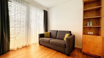 Una sala de estar con un sofá gris, una estantería de madera y un ventanal con cortinas traslúcidas.
