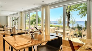 Un restaurant doté de grandes fenêtres donnant sur l’eau et les arbres. Des tables et des chaises sont disposées pour accueillir les clients.