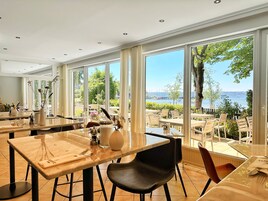 Restaurante com grandes janelas que oferecem vista para a água e árvores. Mesas e cadeiras dispostas para refeições.