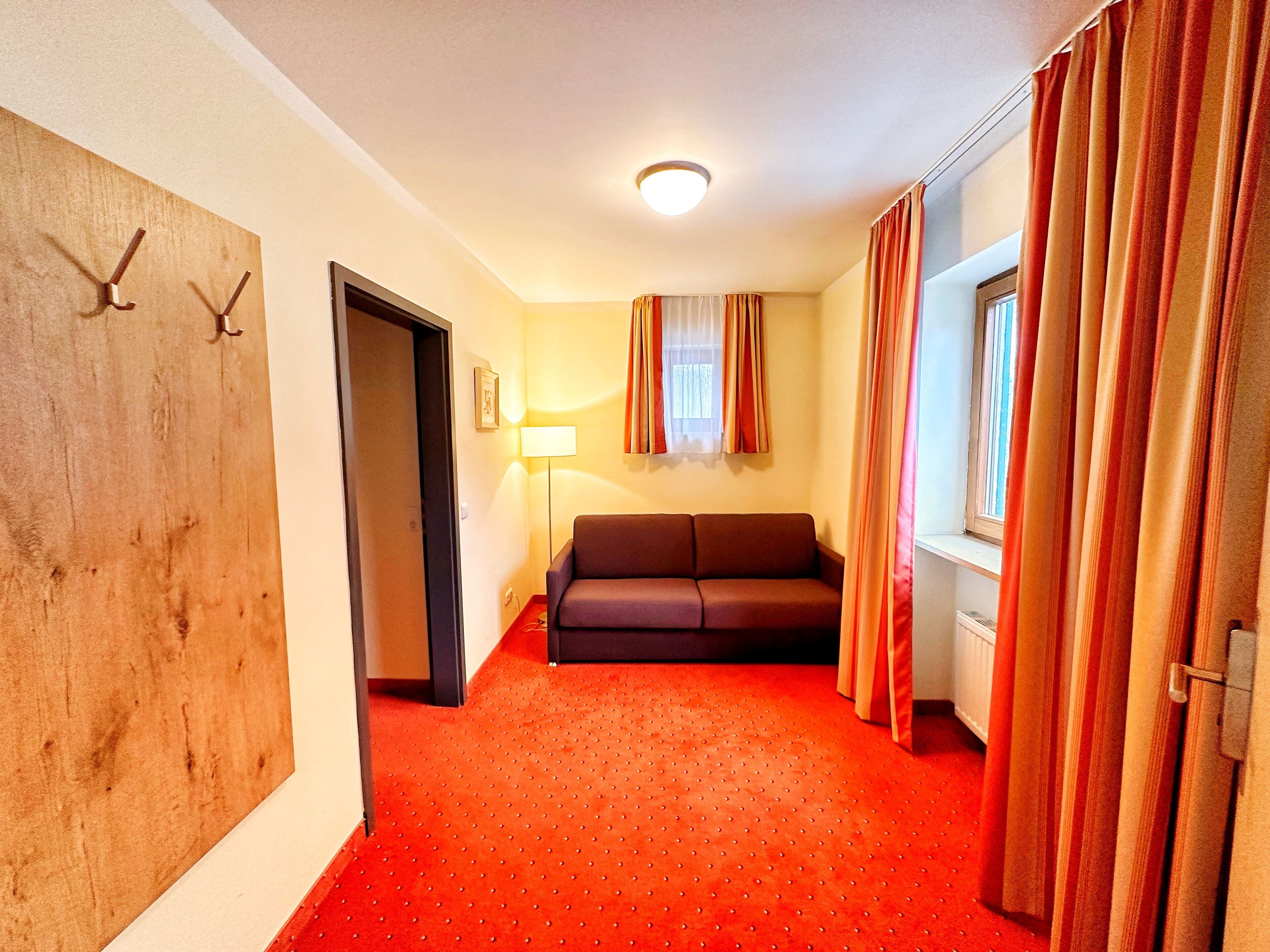 Habitación de hotel con alfombra roja, un sofá, una puerta de madera con ganchos, una lámpara y una ventana con cortinas.