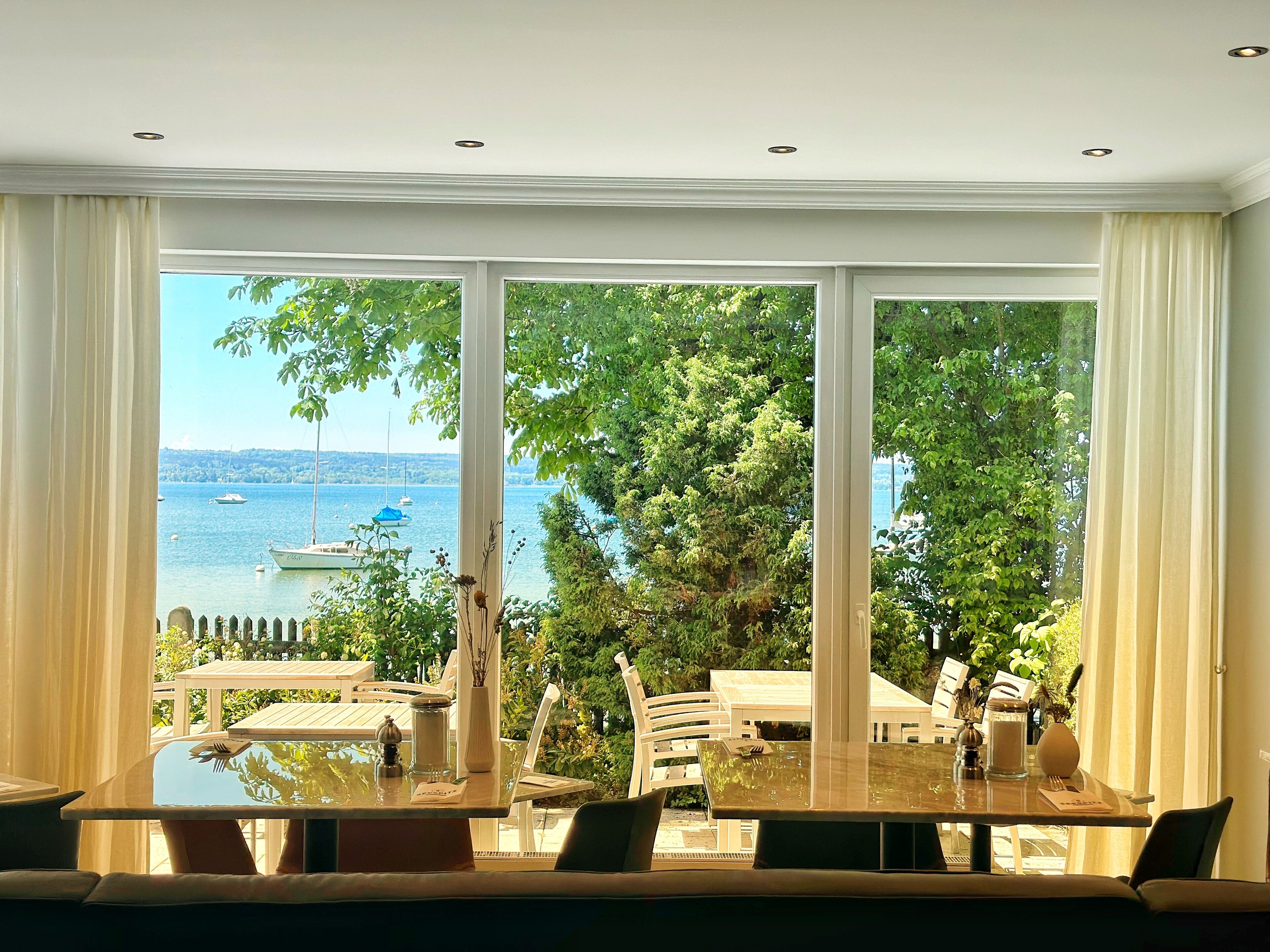 A dining area with a table and chairs, a view of the sea with boats, and greenery outside.