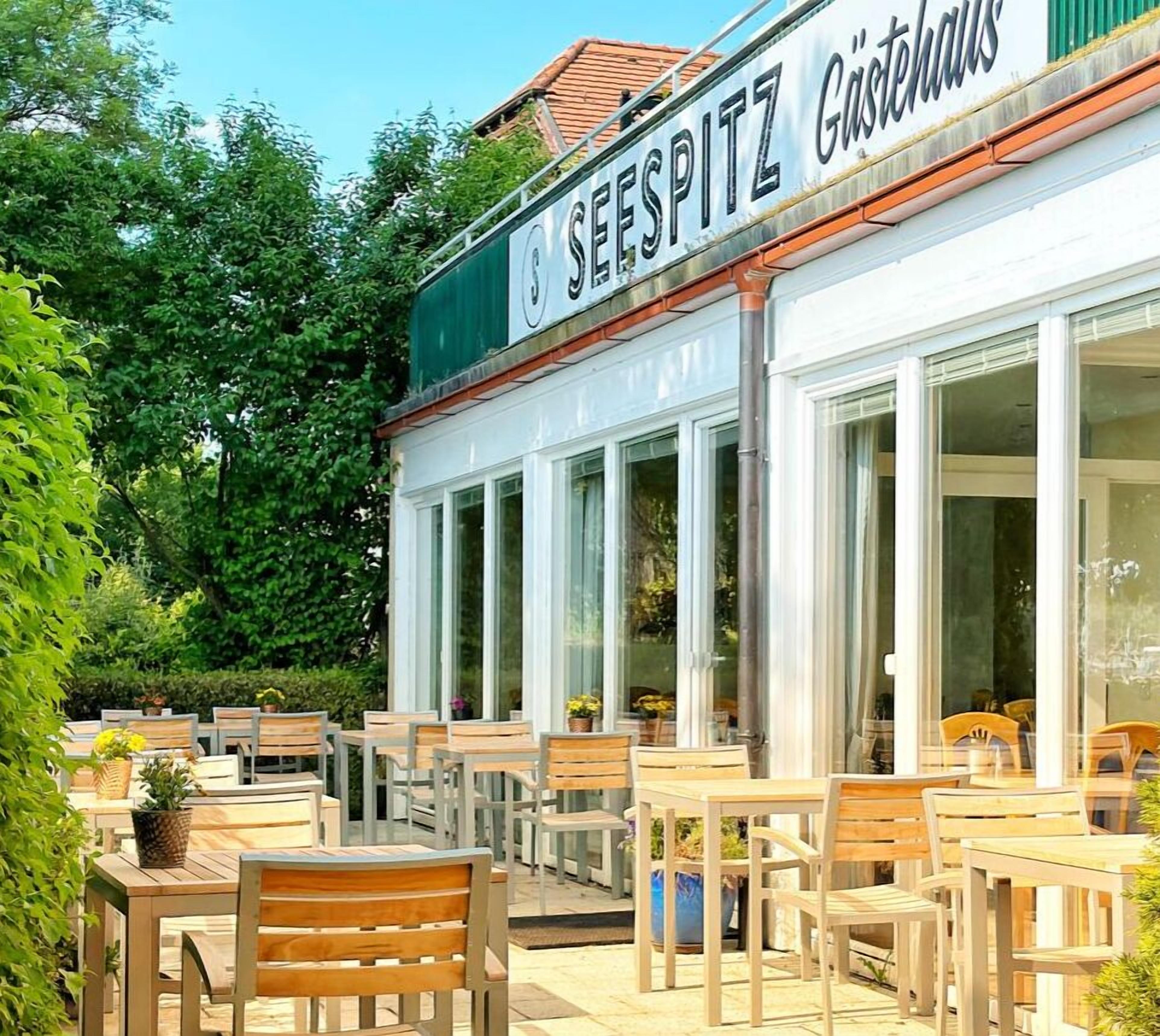 Un área con mesas y sillas de madera, un letrero que dice "SEESPITZ Gasthof" y grandes ventanales.