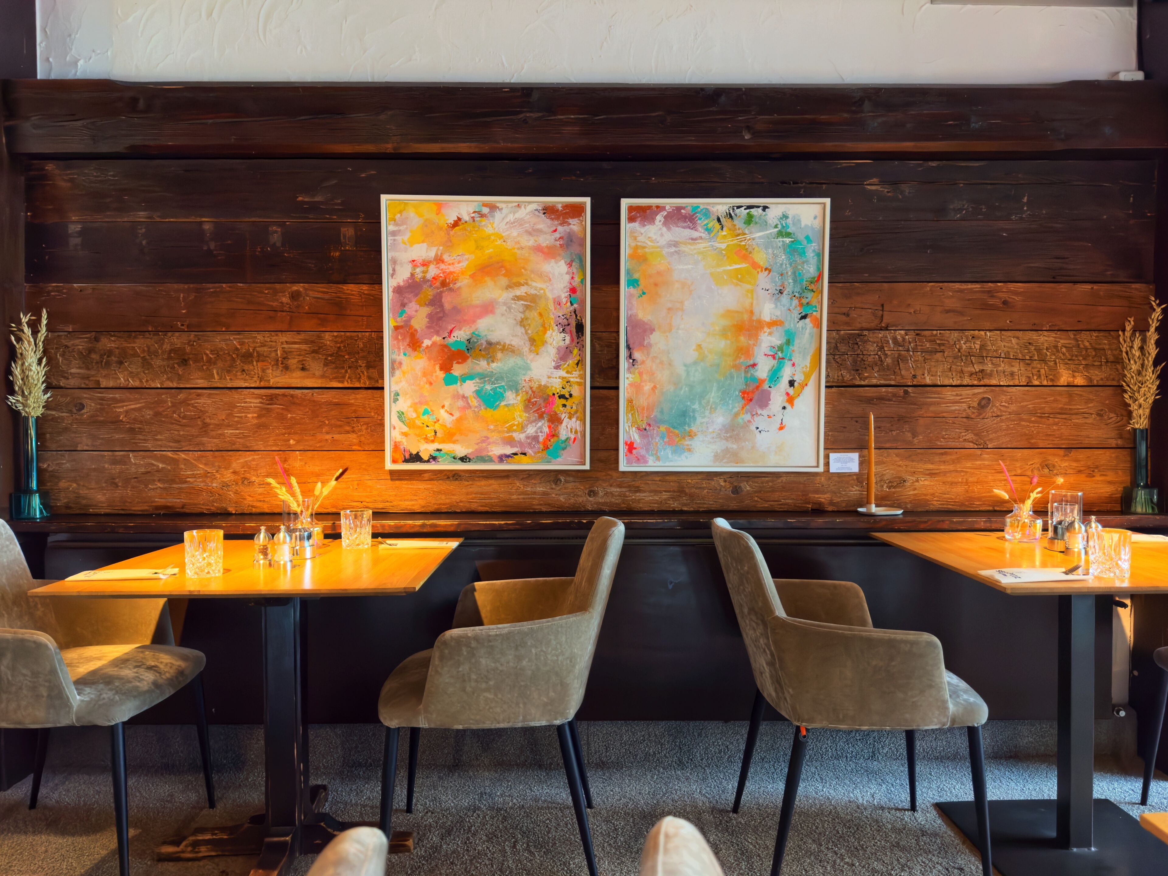 Un restaurant aux murs en bois, orné de deux tableaux abstraits, et dont la table est dressée avec des chaises et des verres.