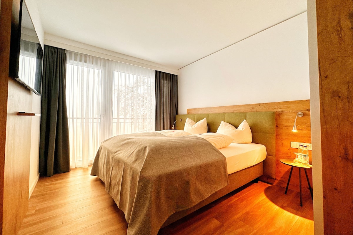 A hotel room with a large bed, a wooden headboard, a bedside table with a lamp, and a large window with curtains.