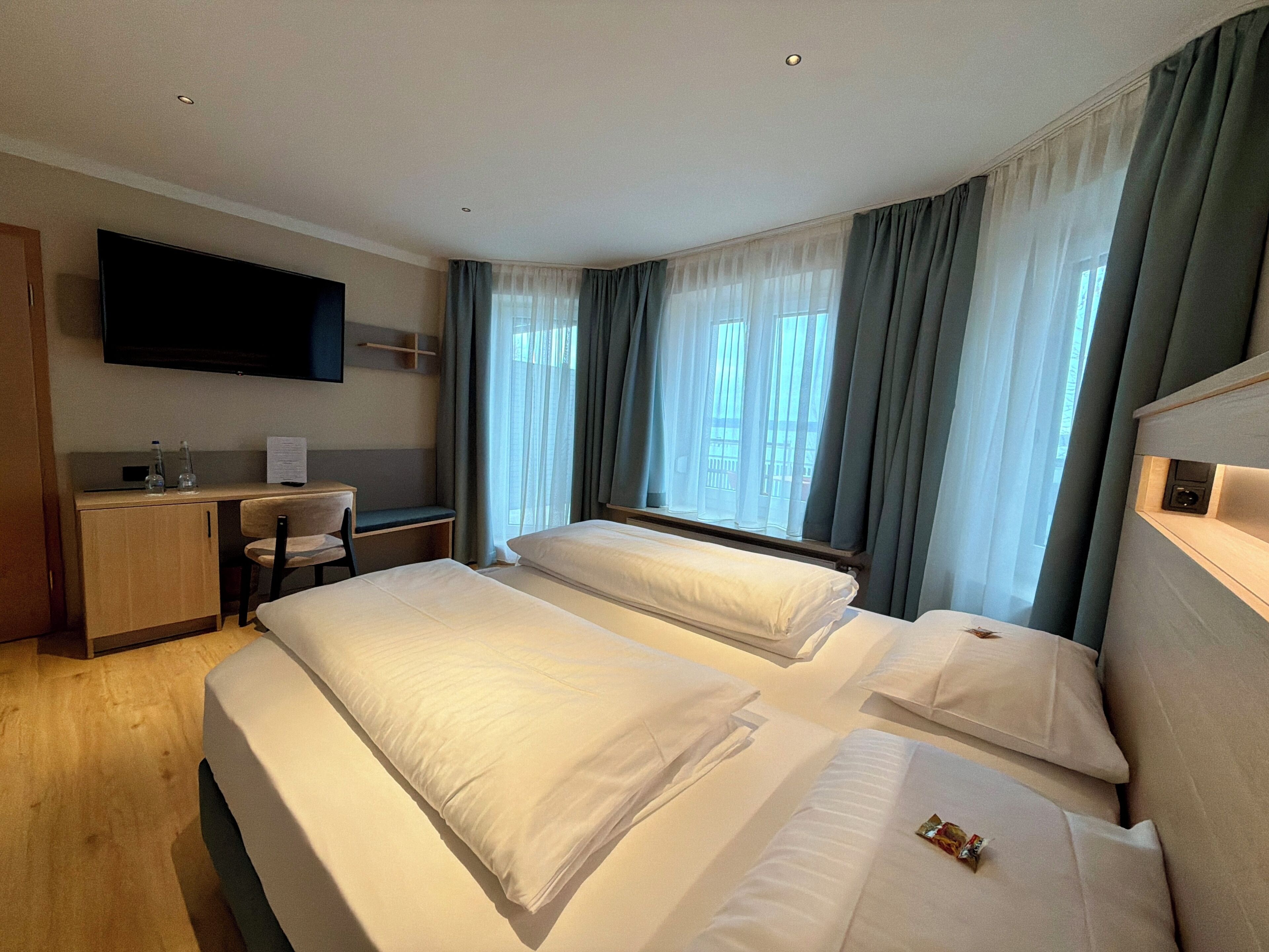 A hotel room with two beds, a desk, a chair, a TV mounted on the wall, and a window with curtains.