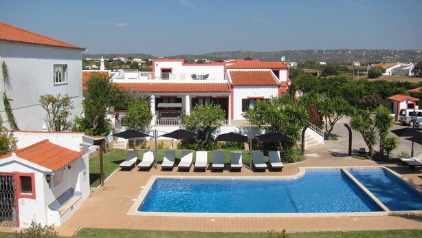 A pool area with lounge chairs and umbrellas, surrounded by buildings with red-tiled roofs and white walls. - Casa da Horta, 1 bedroom apartment, CH1, Pool and Garden (Albufeira)