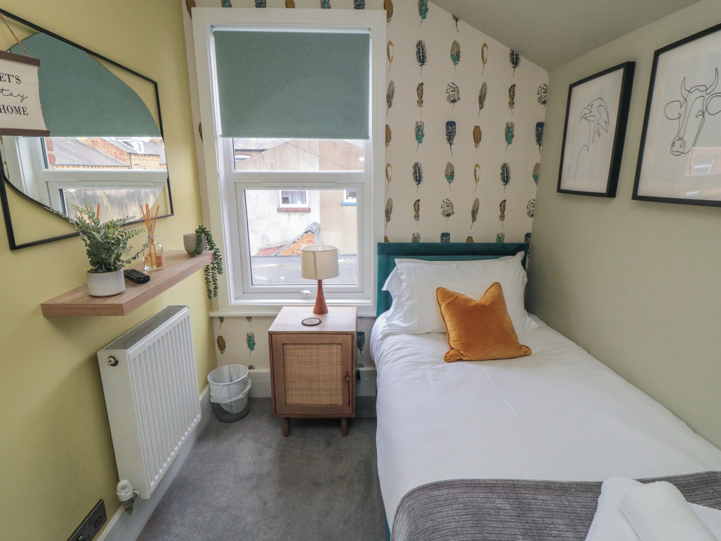 A bedroom with a bed, a nightstand, a radiator, a window, and a wall with feather-patterned wallpaper.