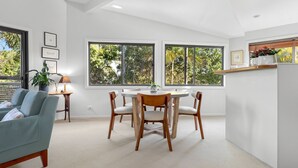 Un'area pranzo moderna con un tavolo rotondo e quattro sedie, ampie finestre con vista sugli alberi, un divano grigio e un piccolo tavolino con una lampada.