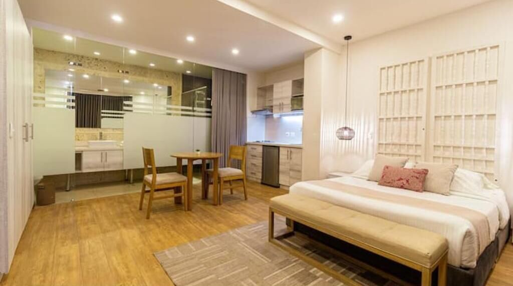 Una habitación de hotel moderna con una cama grande, una mesa de comedor con sillas, una pequeña cocina y un baño visible a través de un panel de vidrio.