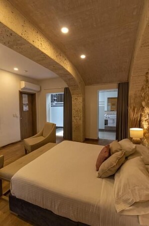 Experience the unique charm of this hotel room with a curved ceiling and exposed brick walls, featuring a large bed, comfortable seating area, and a view of the adjacent kitchen.