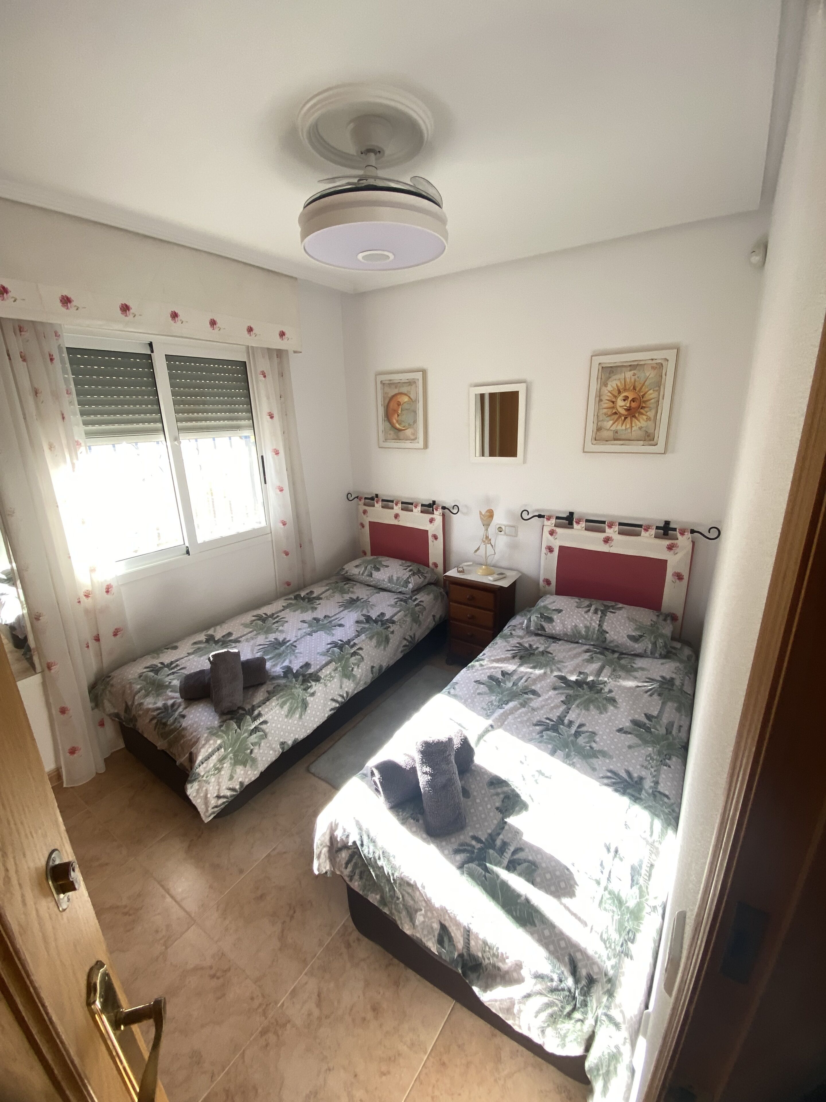Ein Zimmer mit zwei Betten, einem Deckenventilator, einem Fenster mit Jalousien und einem kleinen Tisch mit Lampe.