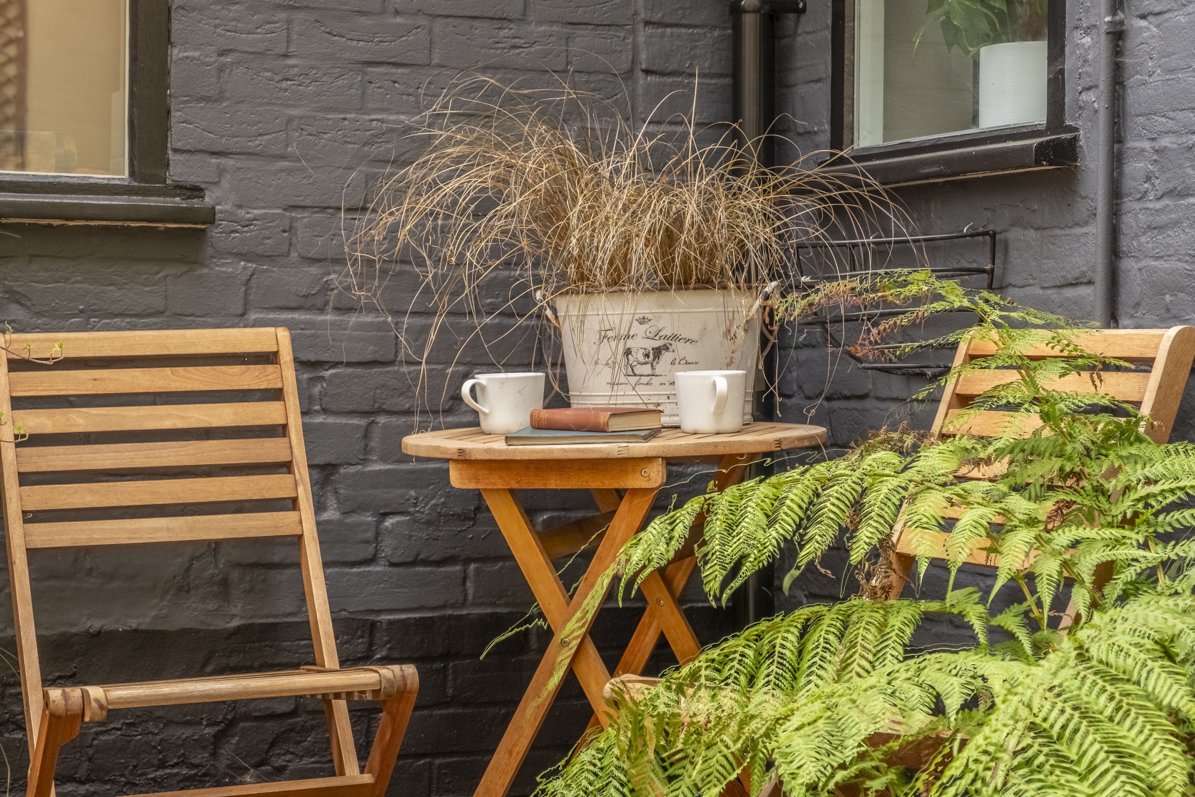 A wooden chair and table set against a dark brick wall, with a potted plant and two white cups on the table.