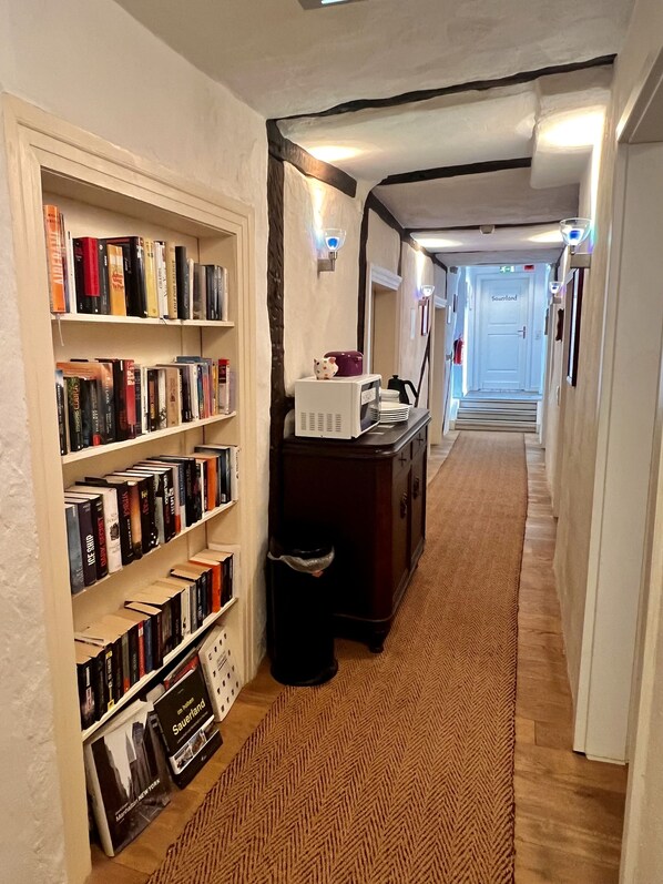 A hallway with bookshelves, a trash bin, a desk with a lamp, and a door labeled "Sauerland". - Hotel Pemü (Arnsberg)