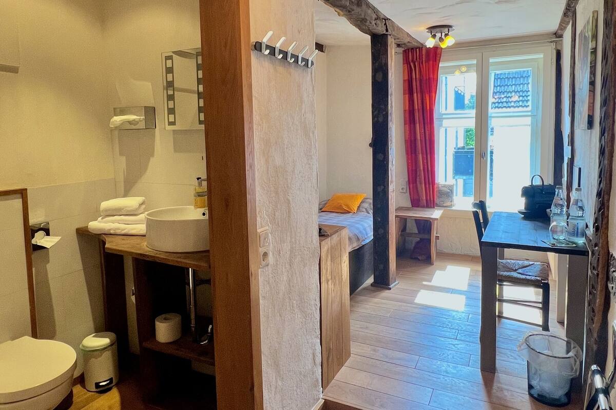 Casa de banho com toucador de madeira, sanita e lavatório. Há um espelho sobre o lavatório e um porta-toalhas. A casa de banho dá acesso a uma área de refeições com mesa e cadeiras.