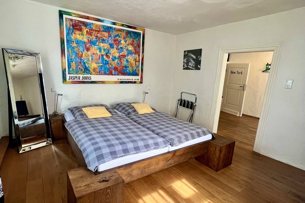 Quarto com uma cama grande, cabeceira de madeira, um quadro, um espelho e uma porta com a inscrição 'New York'.