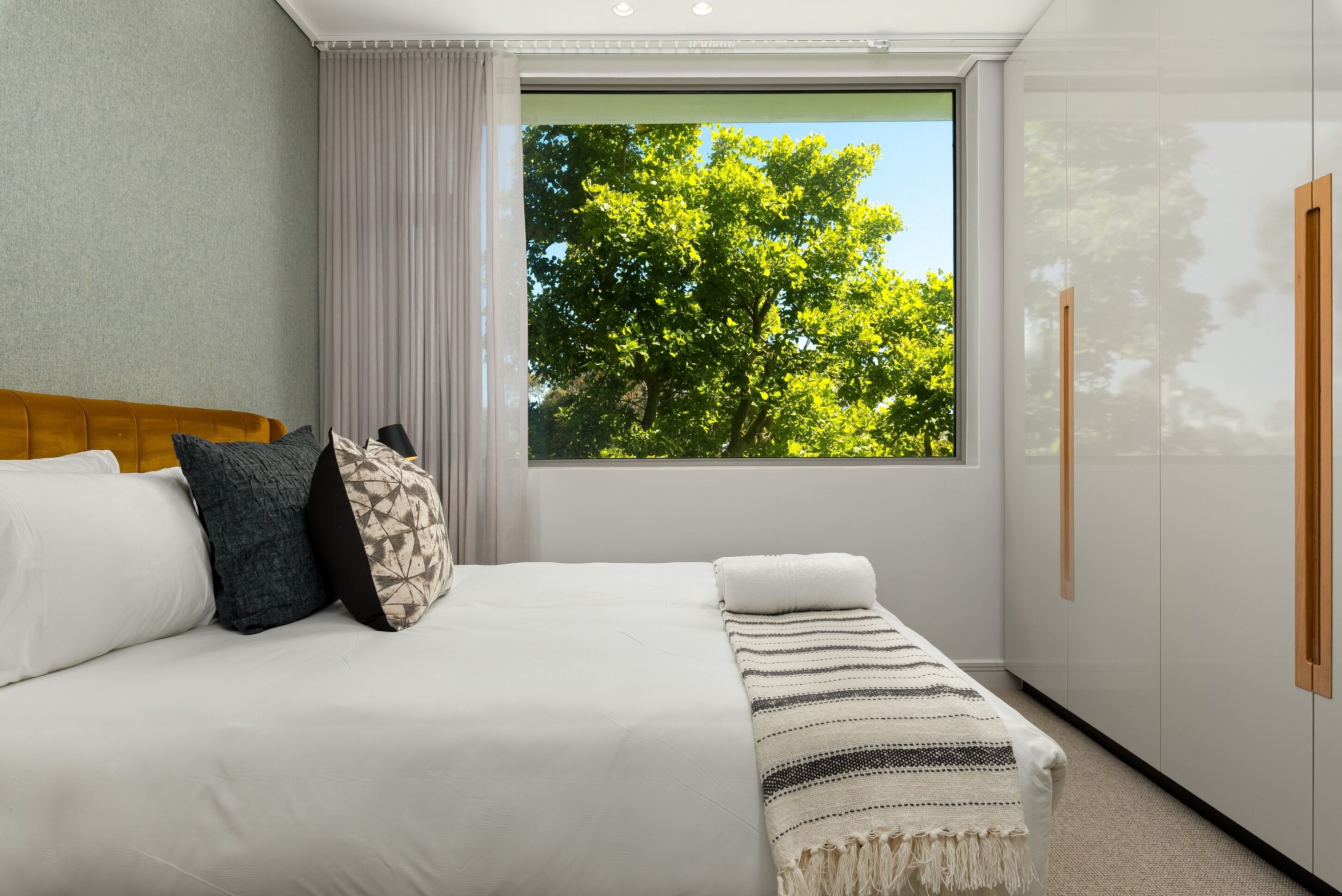 Une chambre d’hôtel avec une grande fenêtre, un lit recouvert de draps blancs, une tête de lit en bois et une vue sur les arbres à l’extérieur.