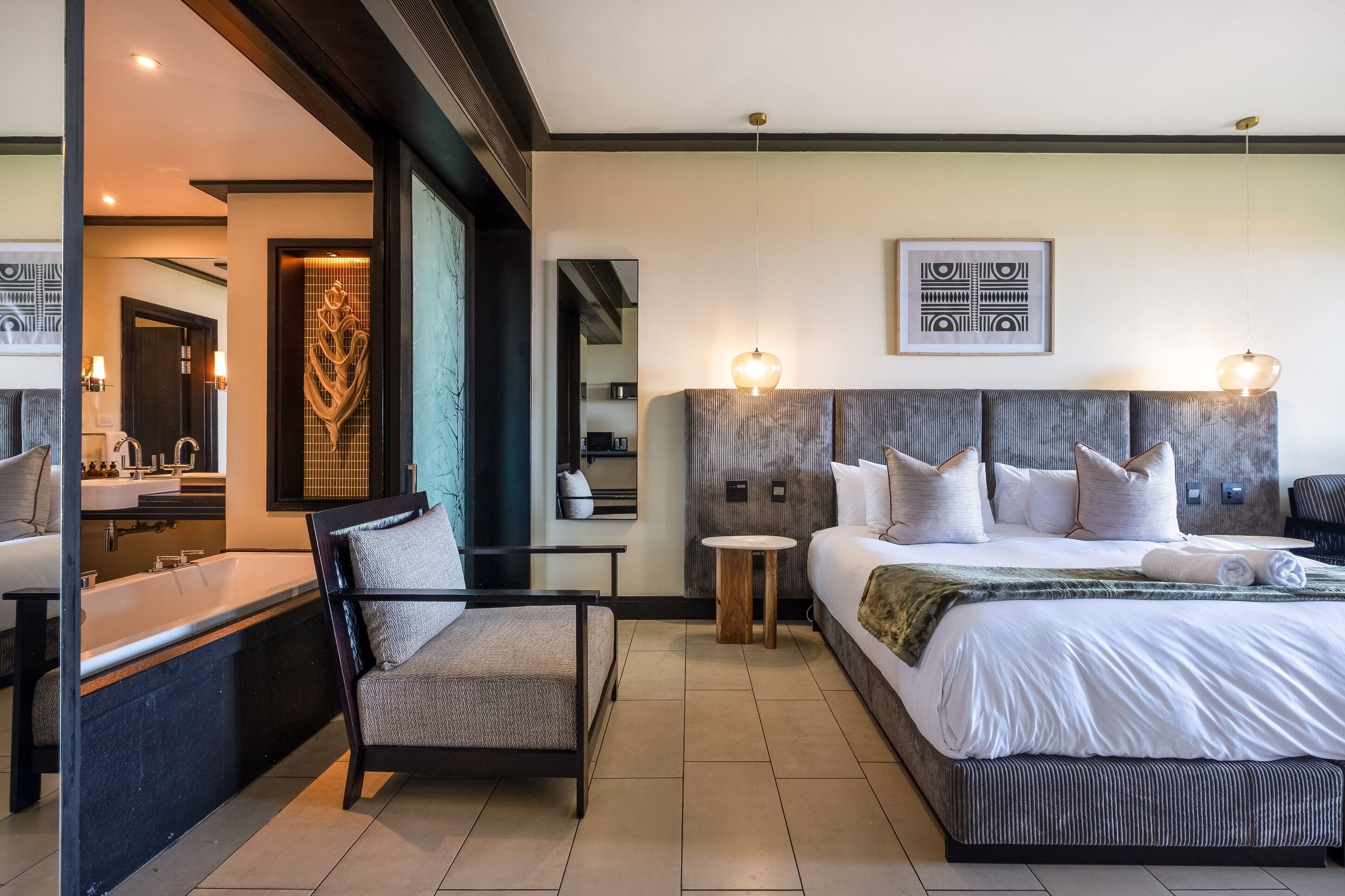 Une chambre d’hôtel moderne dotée d’un grand lit, d’un coin salon avec un fauteuil, d’une petite table ronde et d’une salle de bain avec une baignoire et un lavabo.
