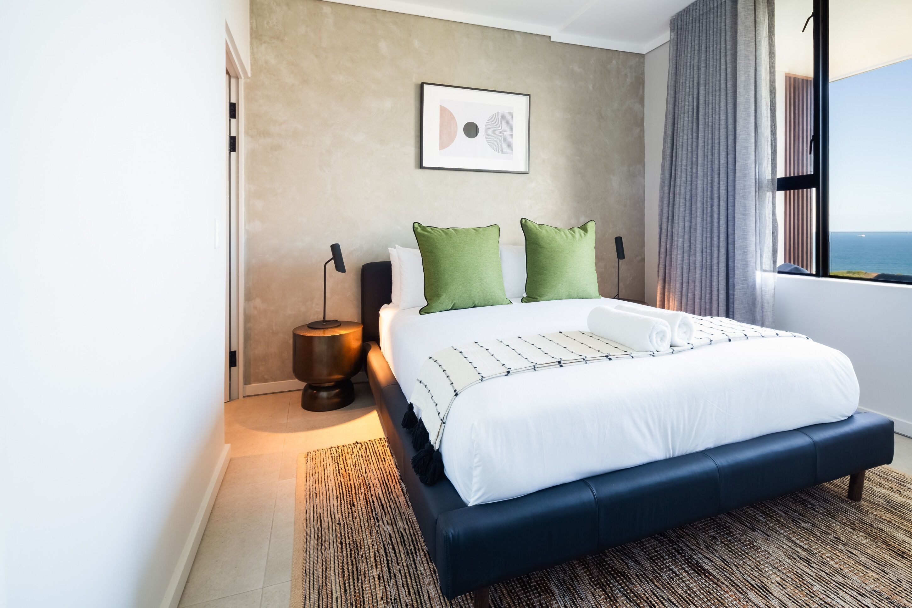 Un dormitorio moderno con una cama grande, almohadas verdes, un cuadro enmarcado en la pared y vistas al mar a través de una ventana.