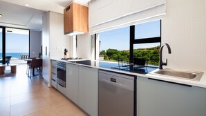 Une cuisine moderne dotée d’un grand îlot central, d’un four encastré et d’un lave-vaisselle. Elle comprend une grande fenêtre offrant une vue sur la verdure, ainsi qu’un coin repas avec une table et des chaises.