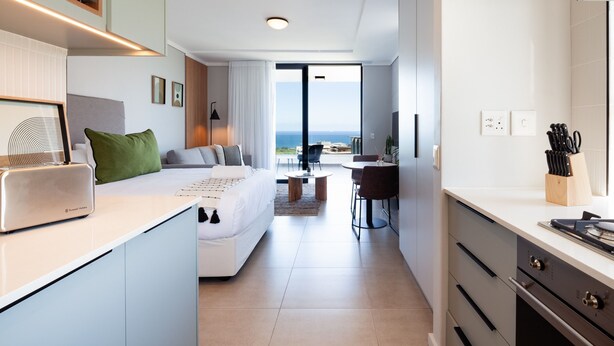 Une chambre d’hôtel moderne dotée d’une kitchenette, d’un lit avec vue, d’un coin repas et d’une grande fenêtre donnant sur la mer.