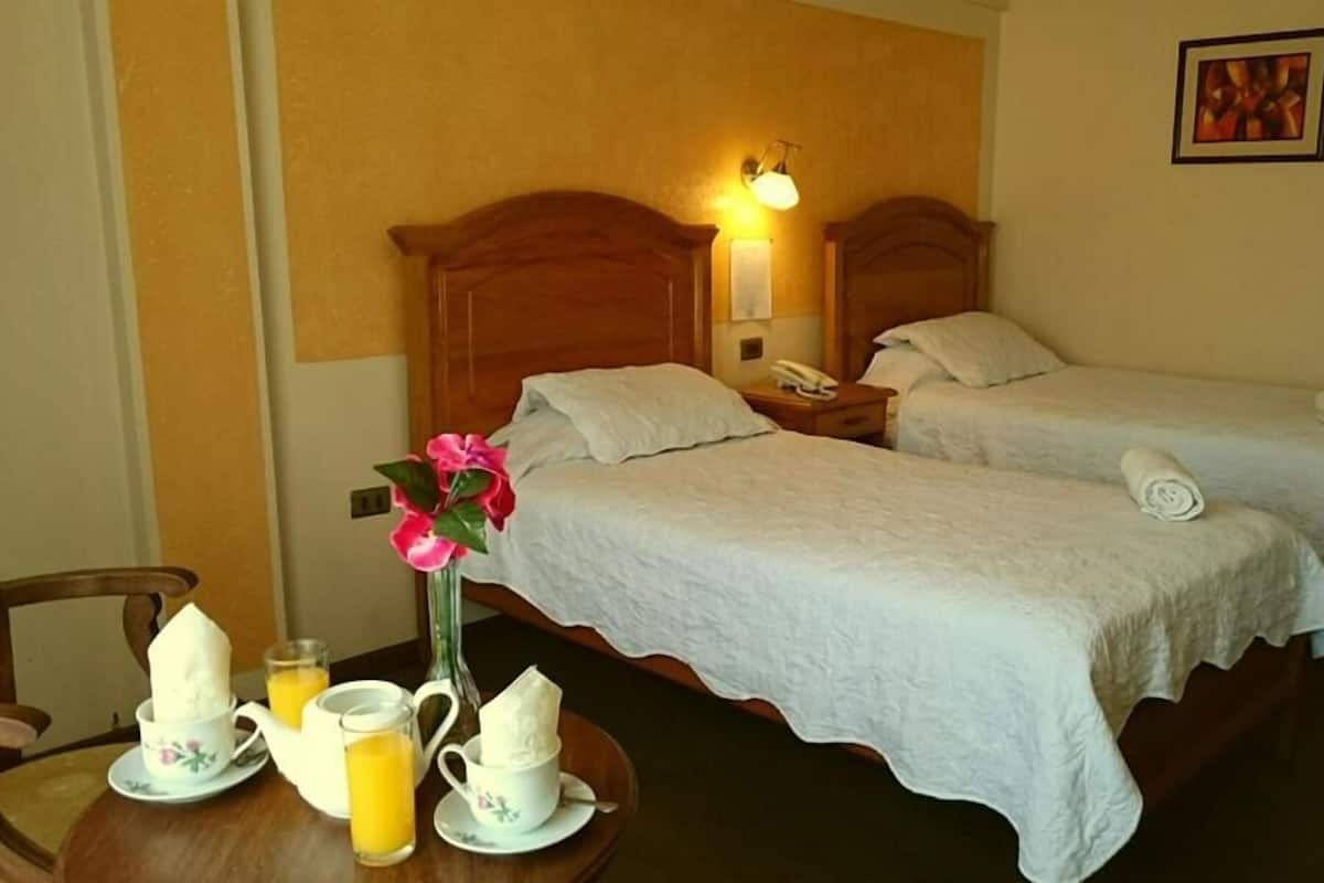 A hotel room with two beds, a bedside table with a lamp, a telephone, and a painting on the wall.