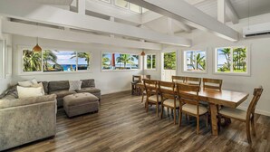 Ein geräumiges Wohnzimmer mit Holzboden, einer Couch, einem Esstisch mit Stühlen und großen Fenstern mit Blick auf eine tropische Landschaft.