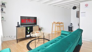 A modern living room with a teal sofa, a black entertainment unit with a TV displaying "NETFLIX," a small round table, and a wicker chair.