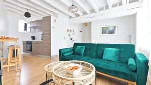 Una sala moderna con un sofá verde, una mesa de centro redonda de mimbre con croissants, un taburete de madera y una cocina al fondo.