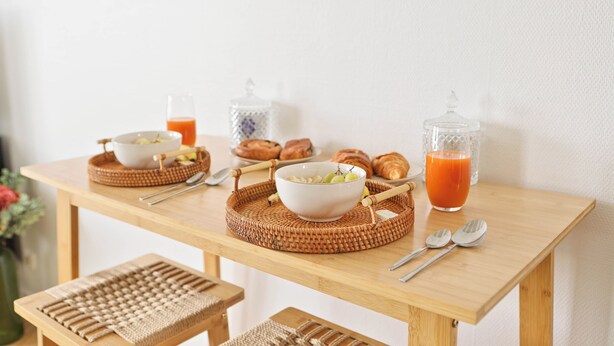Una mesa de madera con individuales tejidos, tazones de comida, croissants y un vaso de jugo de naranja.